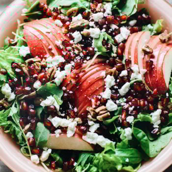 A close up of a bowl of apple slices, pomegranate seeds on a bed of greens.