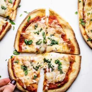 a hand picking up a slice of naan pizza on a white background.