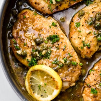 A close up of lemon chicken piccata in a pan with lemon slices and chopped parsley.