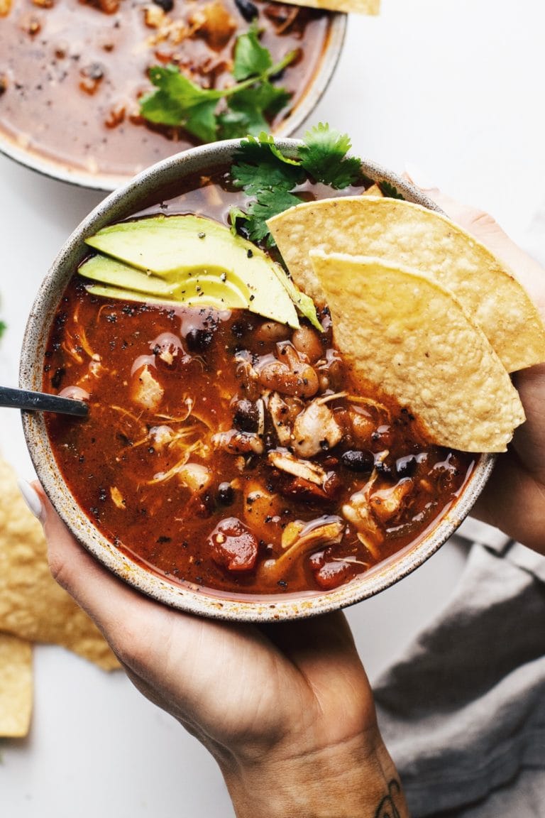 Hands holding a ceramic bowl with slow cooker chicken chili in it with tortilla chips and avocado slices over top.