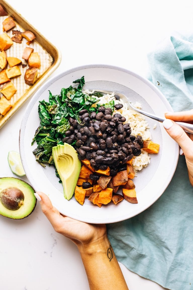Hands holding a grey ceramic bowl with sweet potatoes and black beans in it with a blue napkin and food in the background.