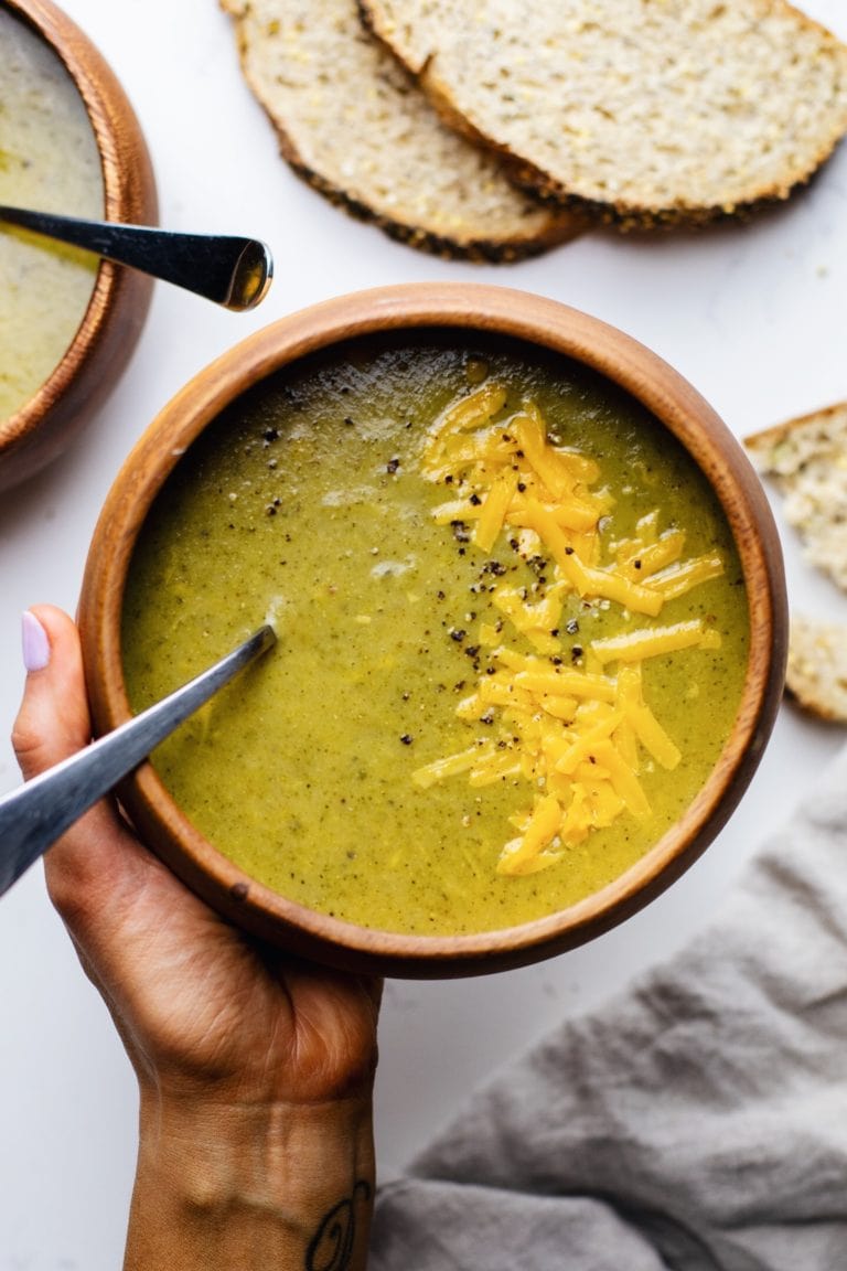 A hand holding a wooden bowl of broccoli and cheese soup with bread slices in the background.
