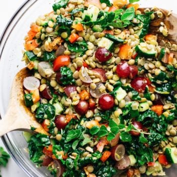 A clear glass bowl with lentil salad and chopped vegetables with 2 wooden spoons in it.