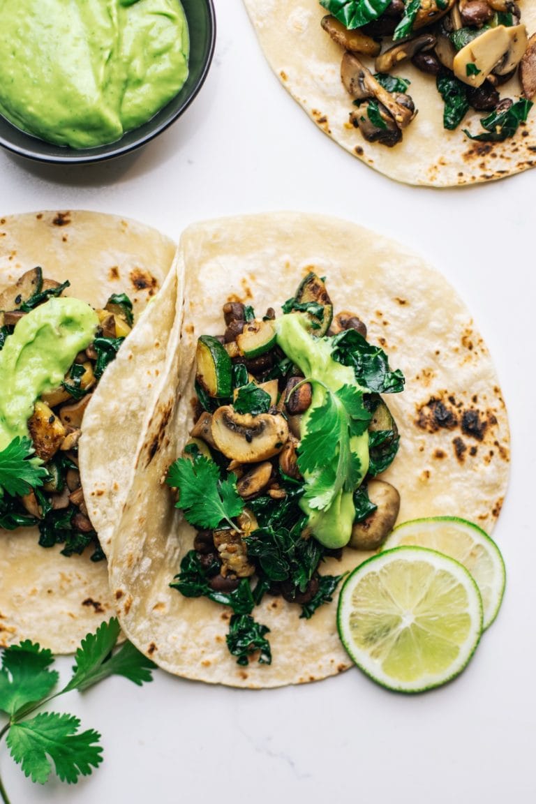 Veggie tacos on a white counter with avocado sauce and lime slices arranged around them.
