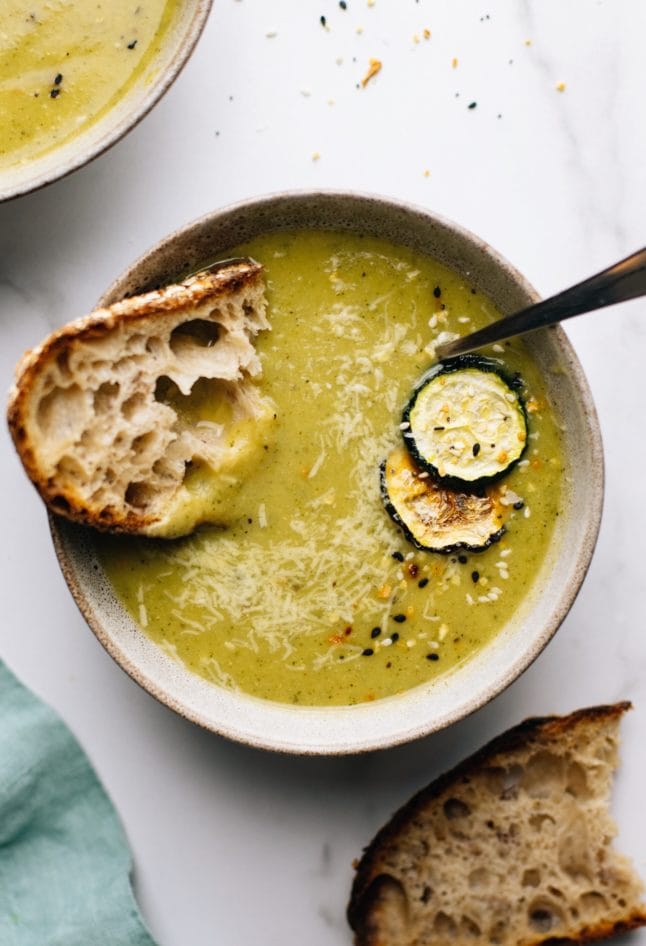 A ceramic bowl on a white counter with zucchini soup in it and a piece of bread in the soup.