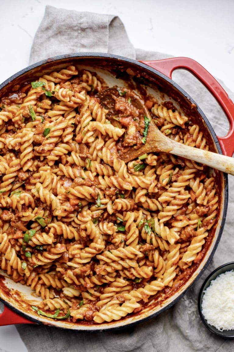 A red pan with pasta noodles and sauce with a wooden spoon in it.