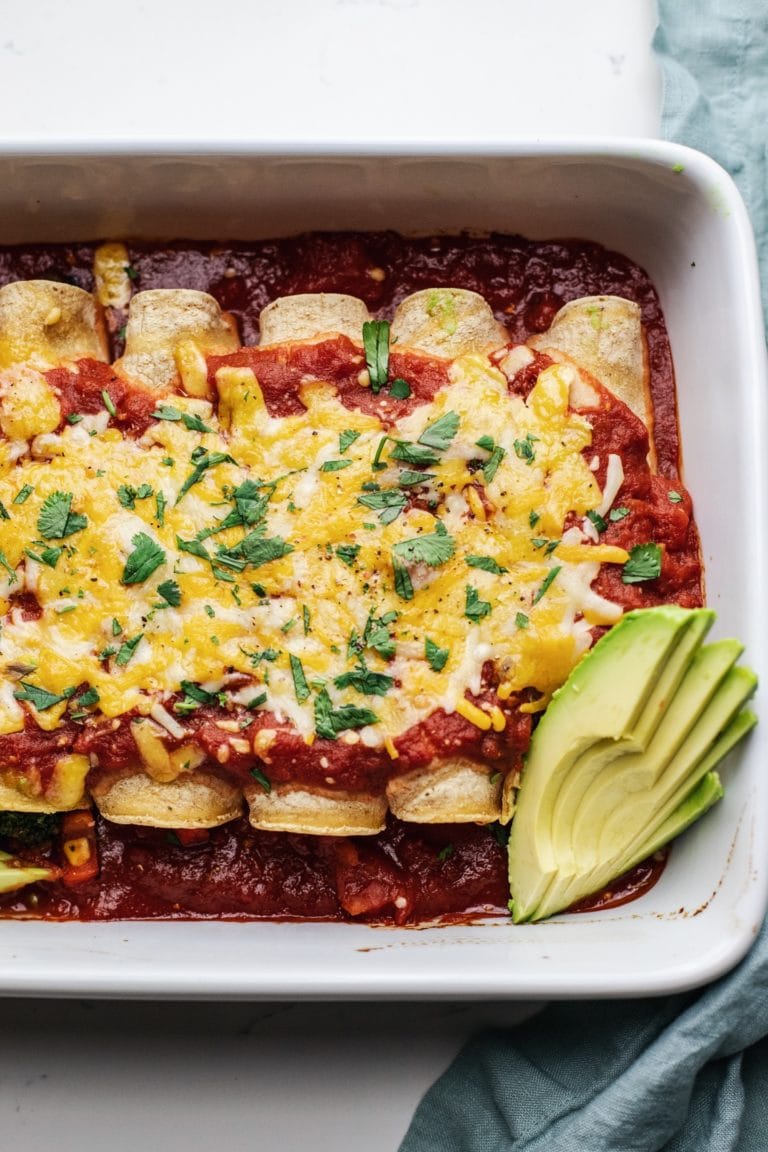 Baked enchiladas in a white dish with avocado slices on the side.