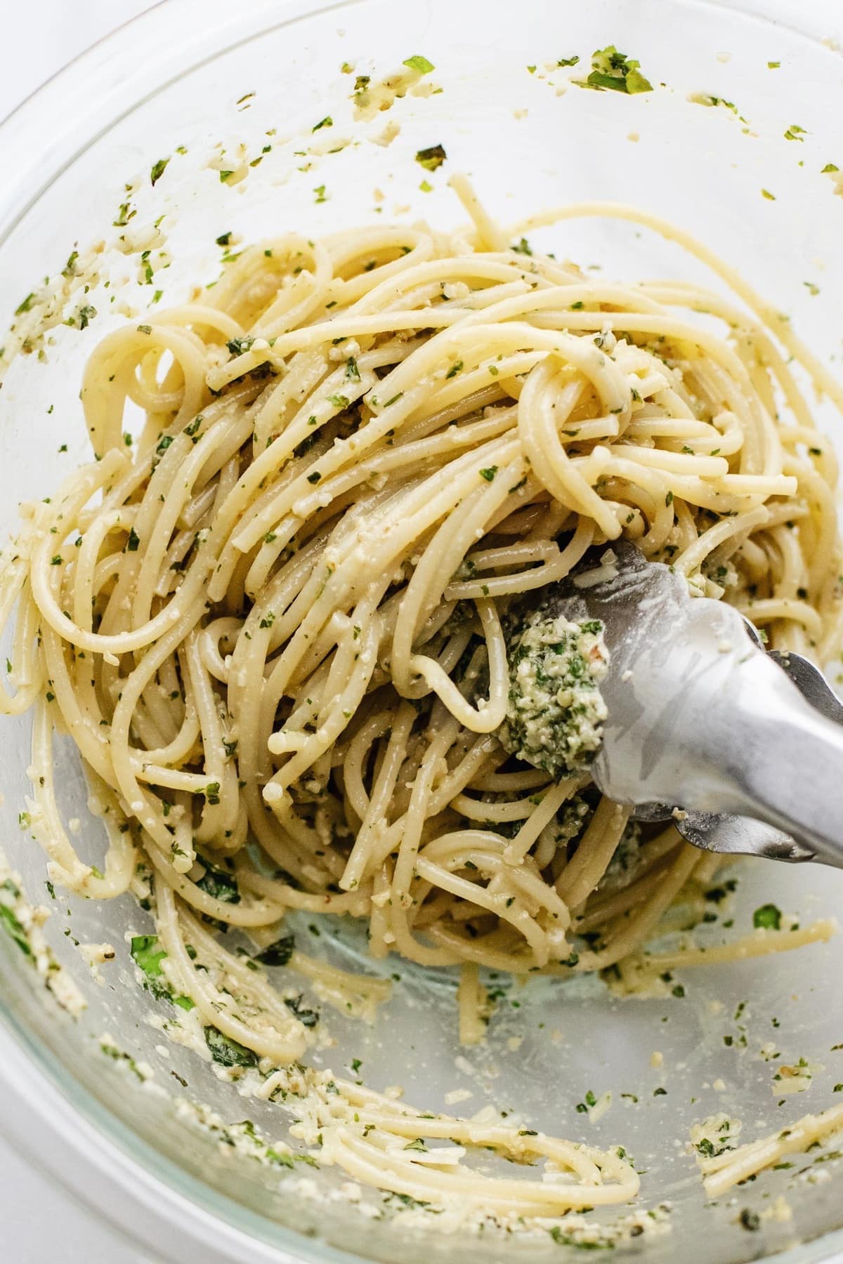 Tongs tossing spaghetti in a clear glass bowl.