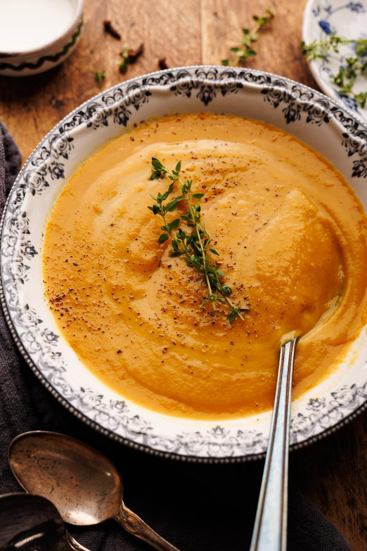 A bowl of sweet potato soup arranged on a wooden board with fresh thyme and a dark napkin with vintage spoons on it placed round the bowl of soup.