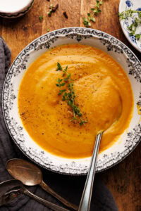 A bowl of sweet potato soup arranged on a wooden board with fresh thyme and a dark napkin with vintage spoons on it placed round the bowl of soup.