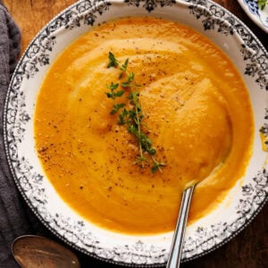 A bowl of sweet potato soup arranged on a wooden board with fresh thyme and a dark napkin with vintage spoons on it placed round the bowl of soup.