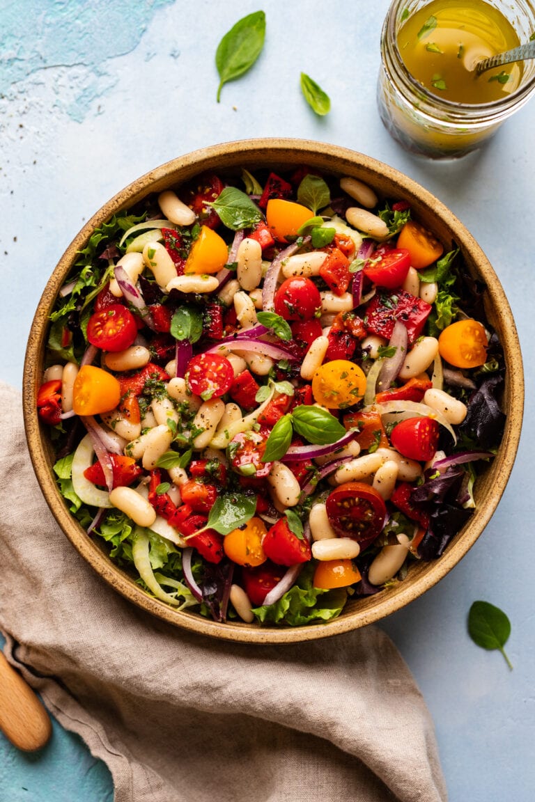 A ceramic bowl with Tuscan cannellini bean salad with assorted vegetables arranged on a light blue counter.