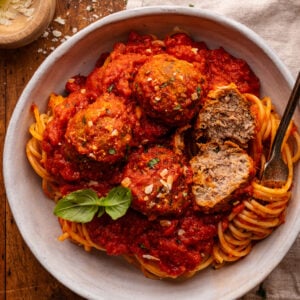 A white bowl with red sauce and Italian meatballs over spaghetti in it. The bowl is sitting on a wooden board with a gray napkin, shredded cheese, and fresh herbs arranged around it.