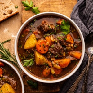 Two white bowls with beef stew and vegetables in it. The bowls are arranged on a wooden cutting board with slices of bread, herbs, and a dark gray napkin placed around the bowls.