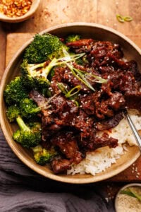 A ceramic bowl with rice topped with beef and broccoli. The bowl is arranged on a wooden board with sesame seeds and a gray napkin placed around it.