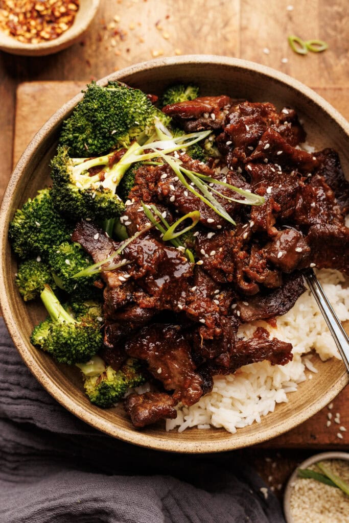 A ceramic bowl with rice topped with beef and broccoli. The bowl is arranged on a wooden board with sesame seeds and a gray napkin placed around it.