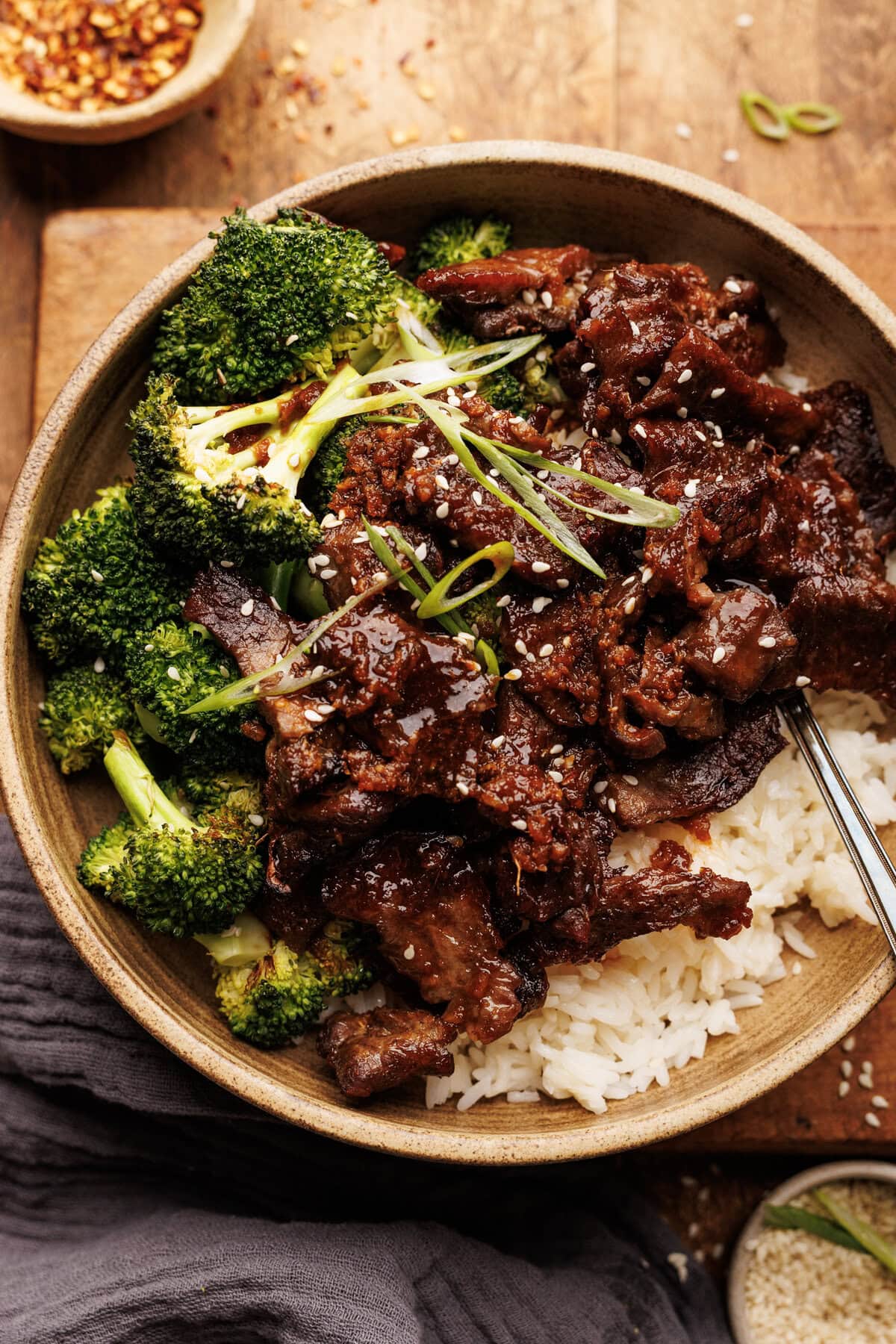 A ceramic bowl with rice topped with beef and broccoli. The bowl is arranged on a wooden board with sesame seeds and a gray napkin placed around it.