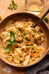 A bowl of shrimp and pasta in it on a tan background with a glass of white wine and herbs decorated around it.