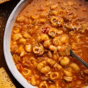 Pasta e fagioli in a white bowl with slices of bread and a parmigiano reggiano rind placed around it on a wooden board.