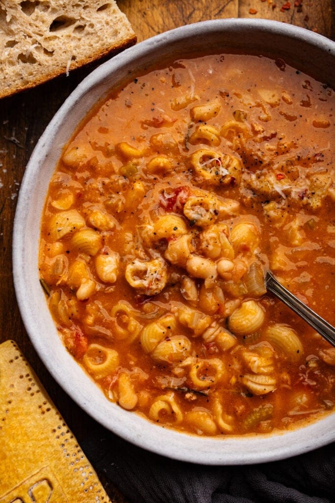 Pasta e fagioli in a white bowl with slices of bread and a parmigiano reggiano rind placed around it on a wooden board.