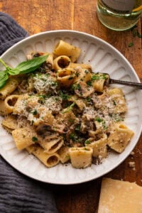 A white bowl with mushroom pasta and cheese sprinkled over the top with a fork in the dish. The bowl of pasta is arranged on a wooden background with a bottle of white wine, a block of parmesan cheese, and a gray napkin placed around it.
