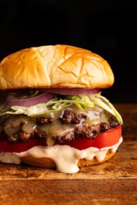 A close up of a smash burger layered on a bun with burger sauce, a tomato and shredded lettuce on a wooden board with a dark background behind it.