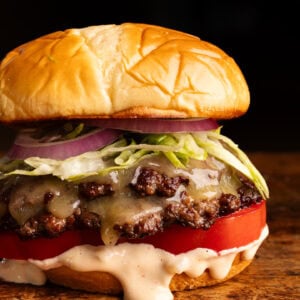 A close up of a smash burger layered on a bun with burger sauce, a tomato and shredded lettuce on a wooden board with a dark background behind it.