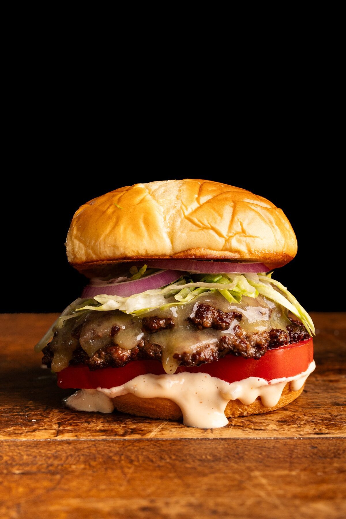 A smash burger layered on a bun with burger sauce, a tomato and shredded lettuce on a wooden board with a dark background behind it.