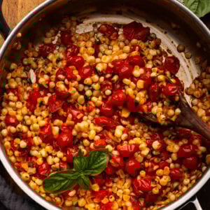 Cooked corn and tomatoes in a stainless steel pan with fresh basil and cherry tomatoes decorated around it.