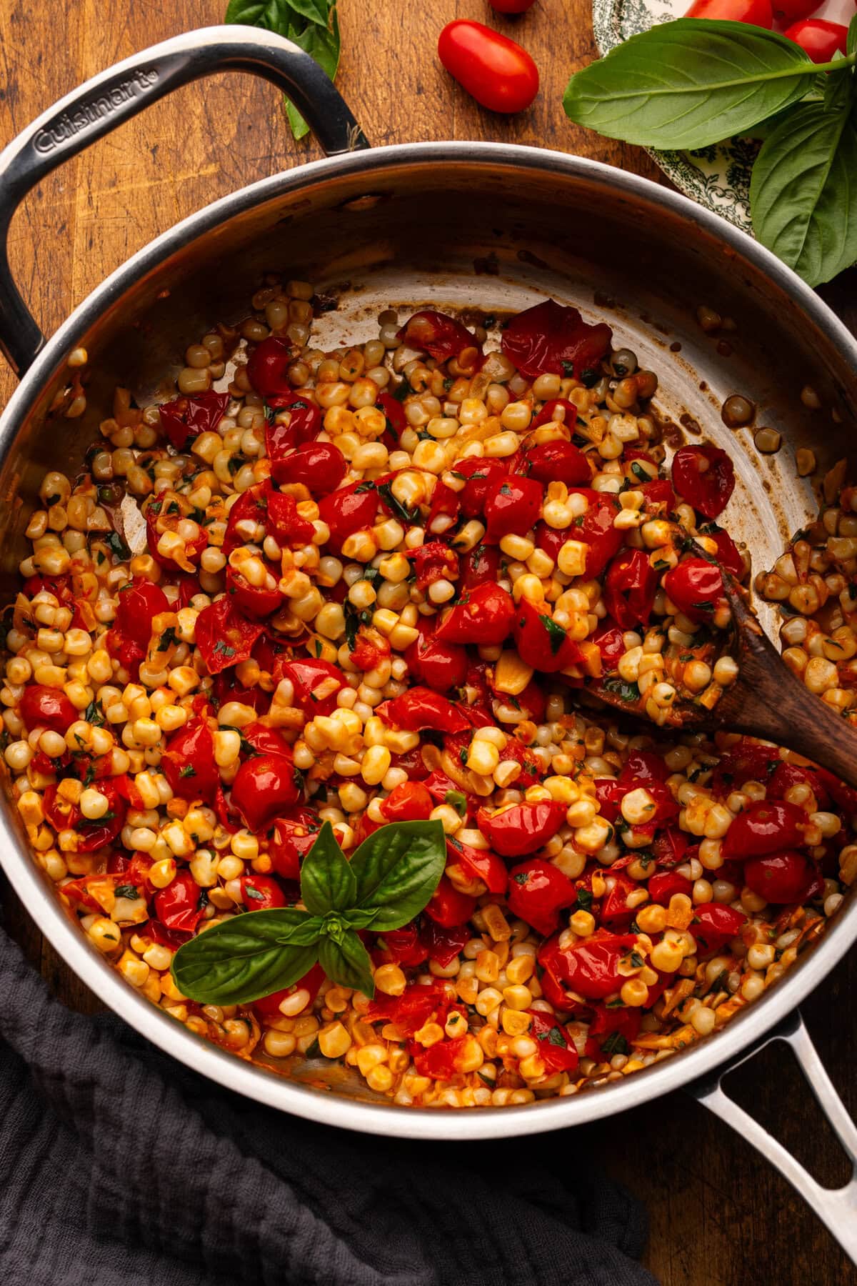 A stainless steel pan with sautéed corn and tomatoes with fresh basil decorated on top. The pan is on a wooden background with a dark napkin and cherry tomatoes and fresh basil placed around the pan.