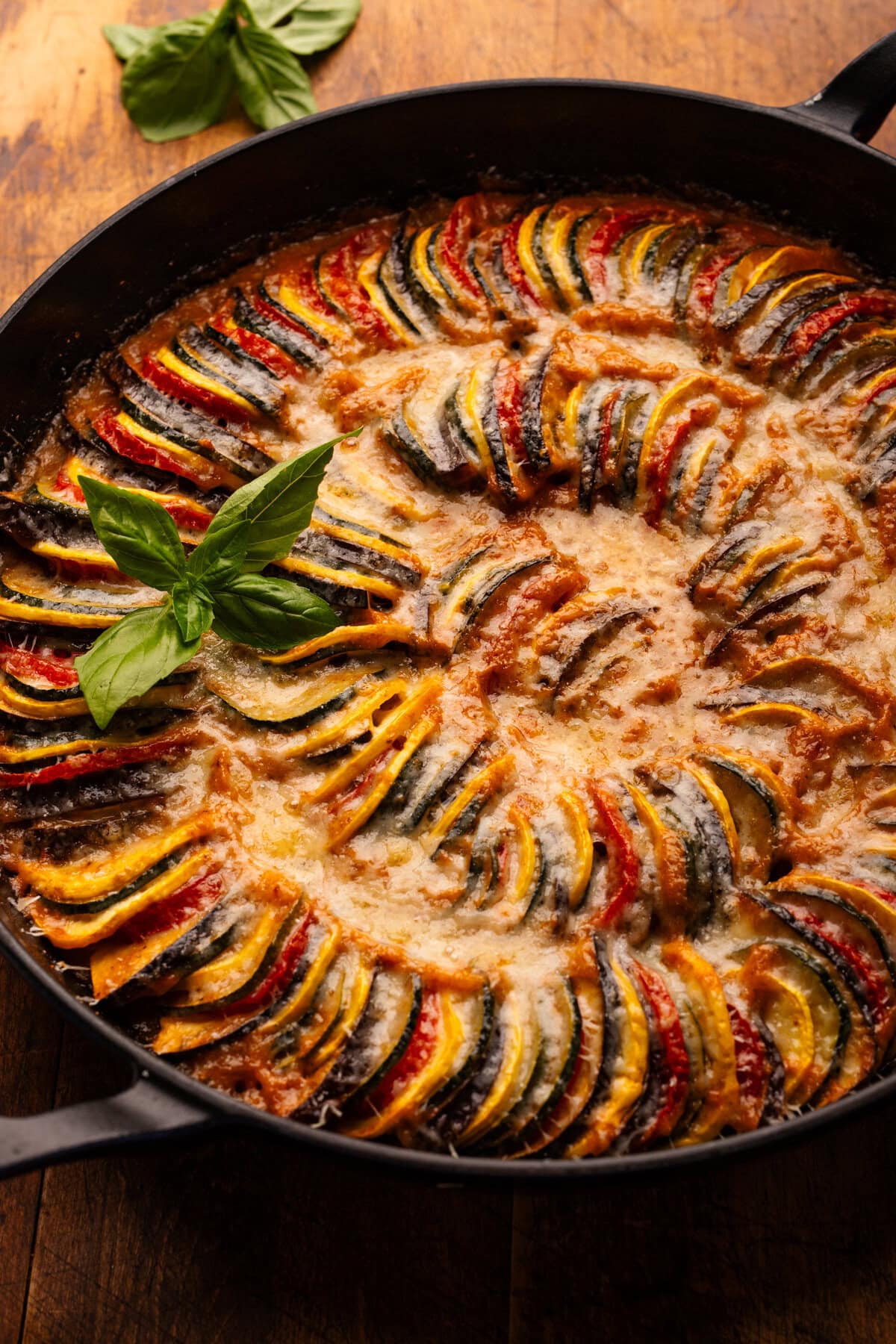 A close up of ratatouille in a cast iron braiser pan on a wooden background with fresh basil arranged around it.