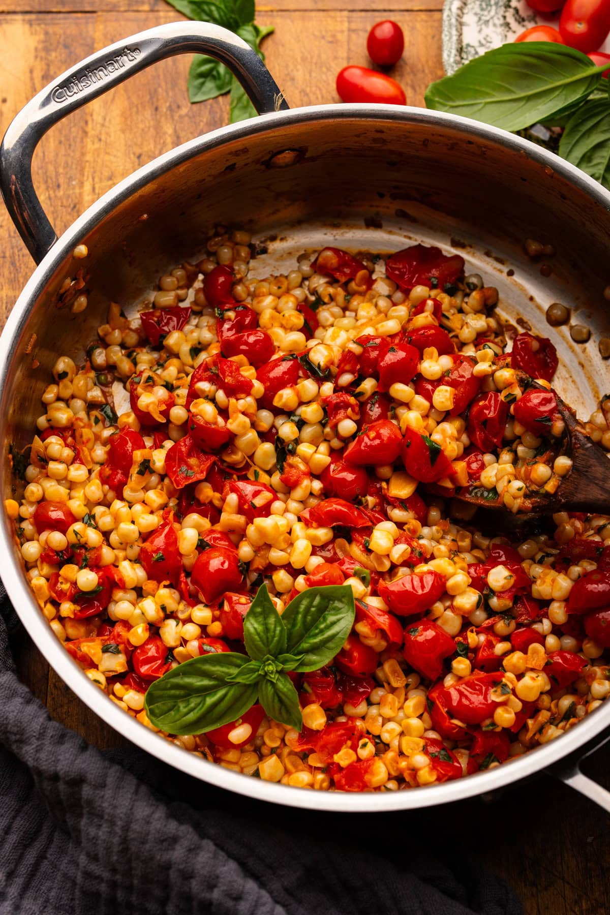 A stainless steel pan with sautéed corn and tomatoes with fresh basil decorated on top. The pan is on a wooden background with a dark napkin and cherry tomatoes and fresh basil placed around the pan.