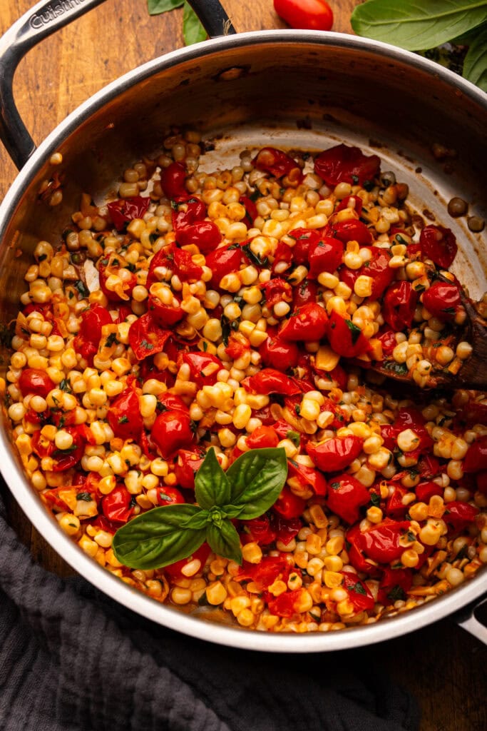 Cooked corn and tomatoes in a stainless steel pan with fresh basil and cherry tomatoes decorated around it.