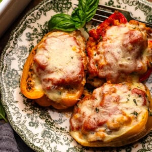 Three stuffed peppers arranged on a green patterned plate that is arranged on a wooden background. Around the plate of stuffed peppers is a dark gray napkin, fresh basil leaves, and a white baking dish with stuffed peppers.