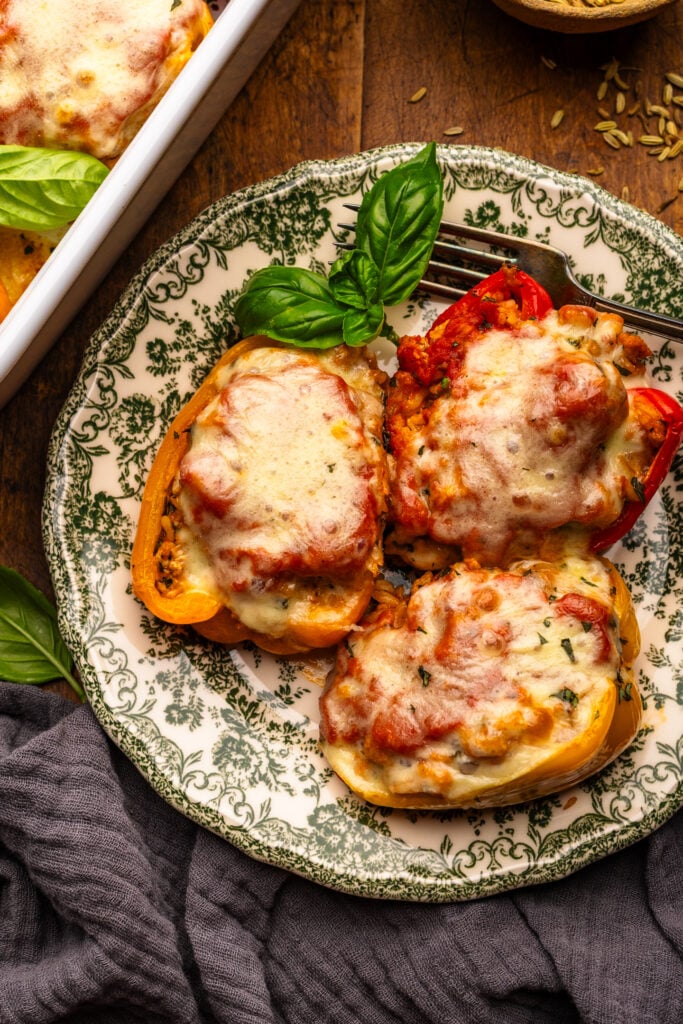 Three stuffed peppers arranged on a green patterned plate that is arranged on a wooden background. Around the plate of stuffed peppers is a dark gray napkin, fresh basil leaves, and a white baking dish with stuffed peppers.