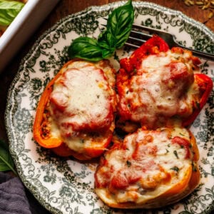 Three stuffed peppers arranged on a green patterned plate that is arranged on a wooden background. Around the plate of stuffed peppers is a dark gray napkin, fresh basil leaves, and a white baking dish with stuffed peppers.