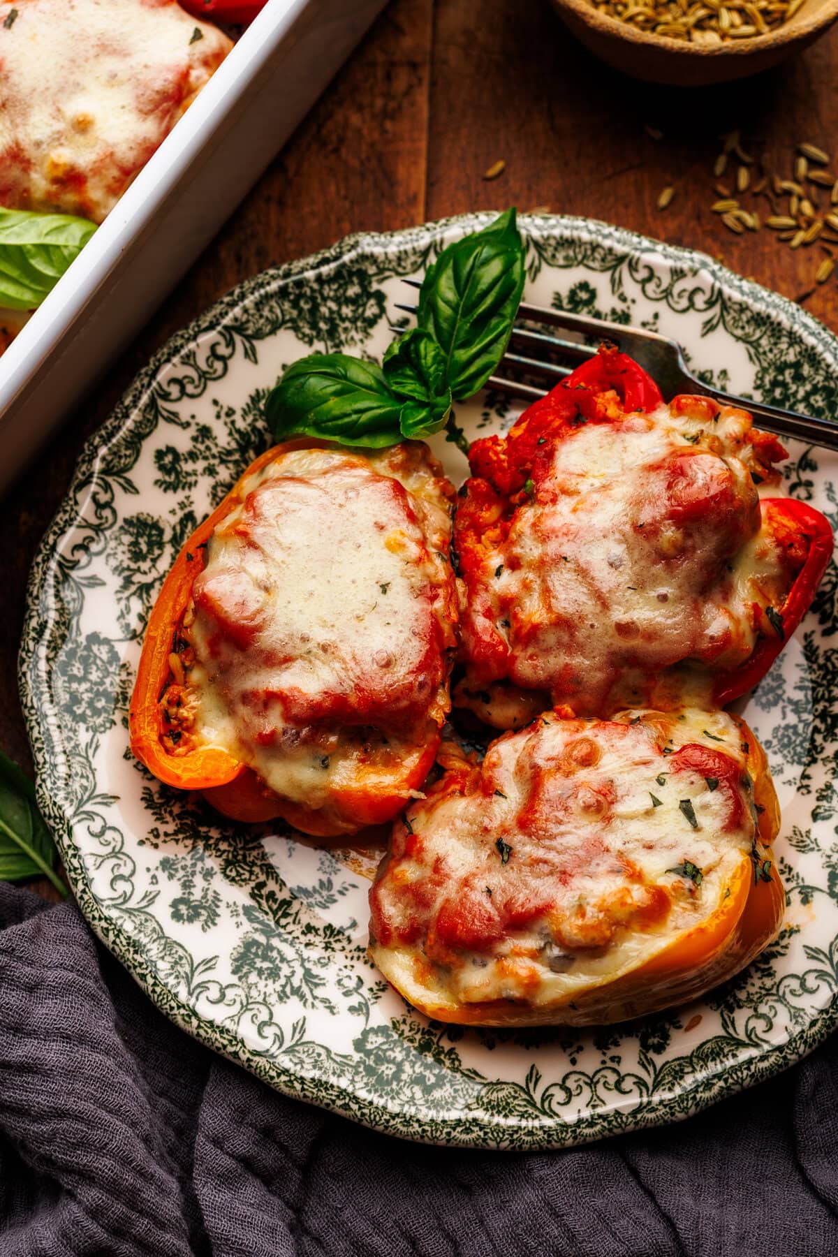 Three stuffed peppers arranged on a green patterned plate that is arranged on a wooden background. Around the plate of stuffed peppers is a dark gray napkin, fresh basil leaves, and a white baking dish with stuffed peppers.