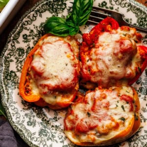 Three stuffed peppers arranged on a green patterned plate that is arranged on a wooden background. Around the plate of stuffed peppers is a dark gray napkin, fresh basil leaves, and a white baking dish with stuffed peppers.