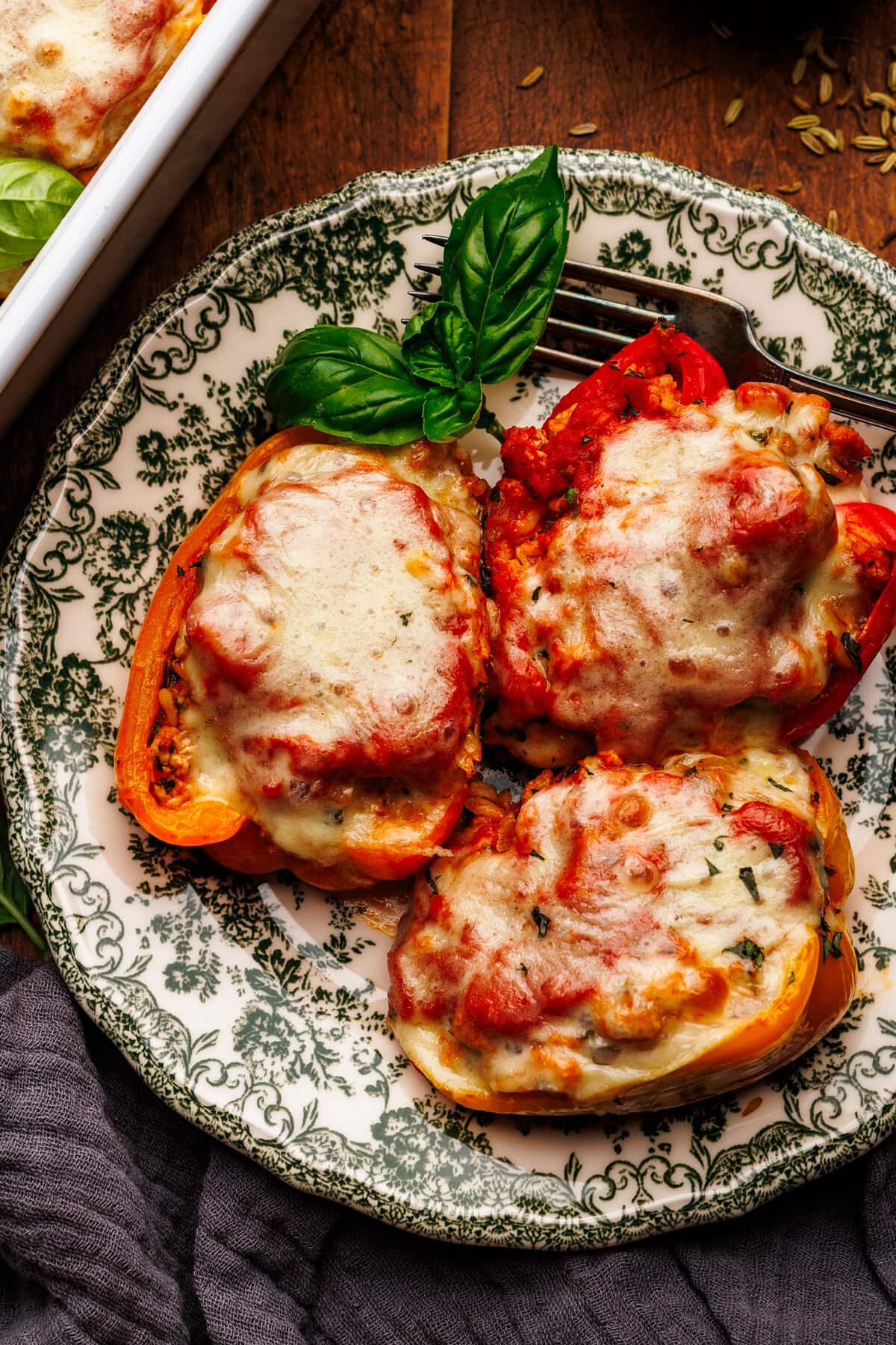 Three stuffed peppers arranged on a green patterned plate that is arranged on a wooden background. Around the plate of stuffed peppers is a dark gray napkin, fresh basil leaves, and a white baking dish with stuffed peppers.