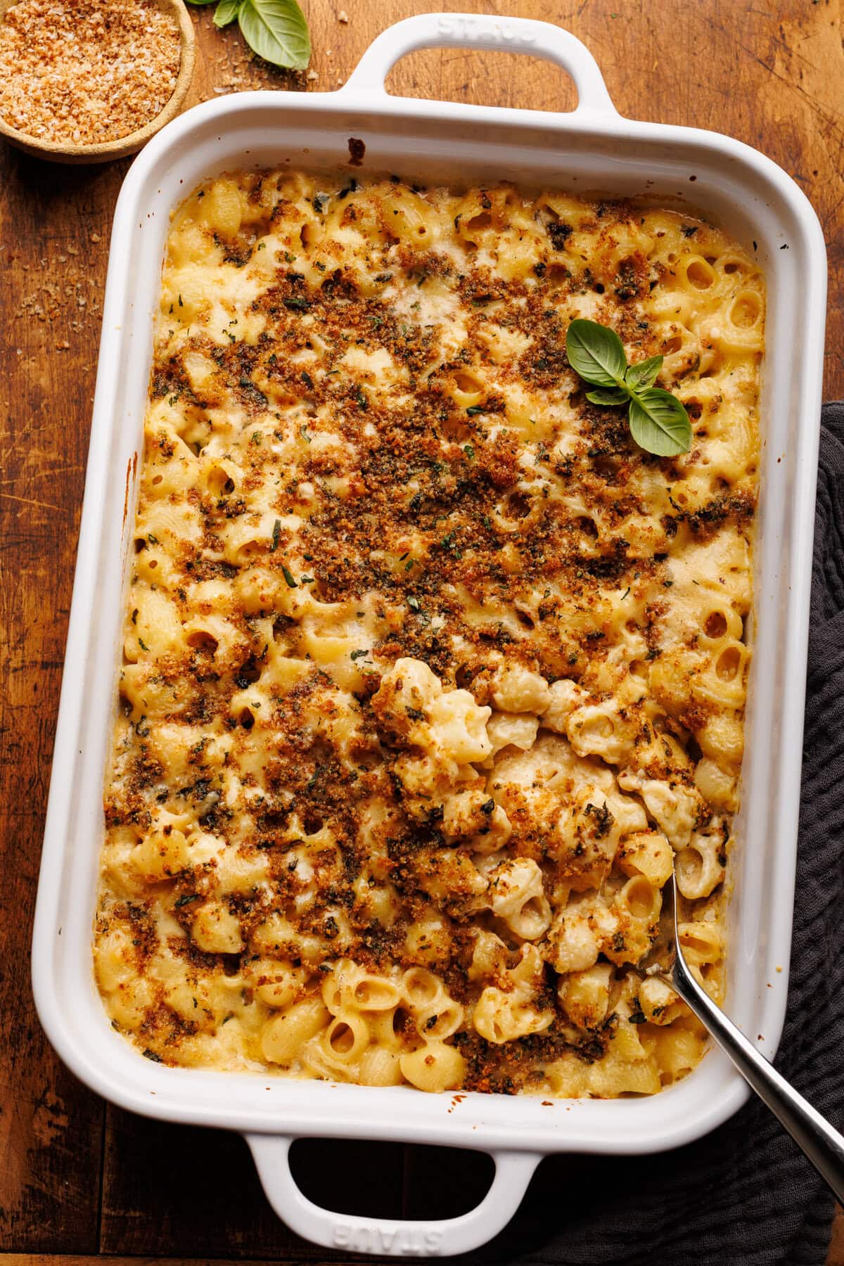 A white glass baking dish with baked mac and cheese in it and bread crumbs on top. There is a silver serving spoon in the mac and cheese. The dish is on a wooden background with a small bowl of breadcrumbs and fresh basil placed around it.
