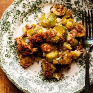 A green vintage plate with Brussels sprout gratin on it with a fork. This plate is arranged on a wooden background with a brown gingham napkin.