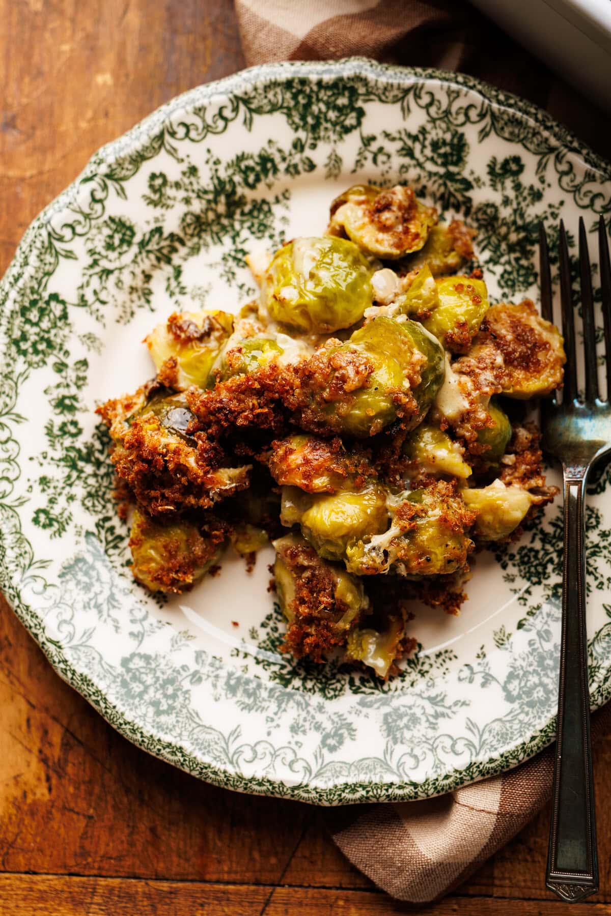A green vintage plate with Brussels sprout gratin on it with a fork. This plate is arranged on a wooden background with a brown gingham napkin.