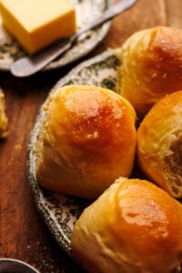 A close up of homemade dinner rolls on a plate with butter on a plate next to them, sitting on a wooden background.