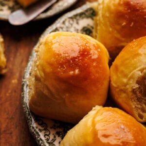 A close up of homemade dinner rolls on a plate with butter on a plate next to them, sitting on a wooden background.