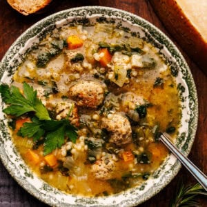 A bowl of Italian wedding soup arranged on a wooden board with slices of bread, a block of Parmigiano Reggiano, and a dark gray napkin placed around it.