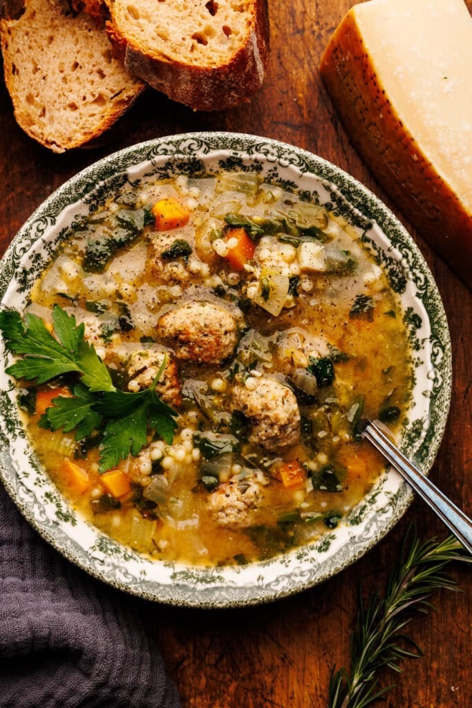 A bowl of Italian wedding soup arranged on a wooden board with slices of bread, a block of Parmigiano Reggiano, and a dark gray napkin placed around it.
