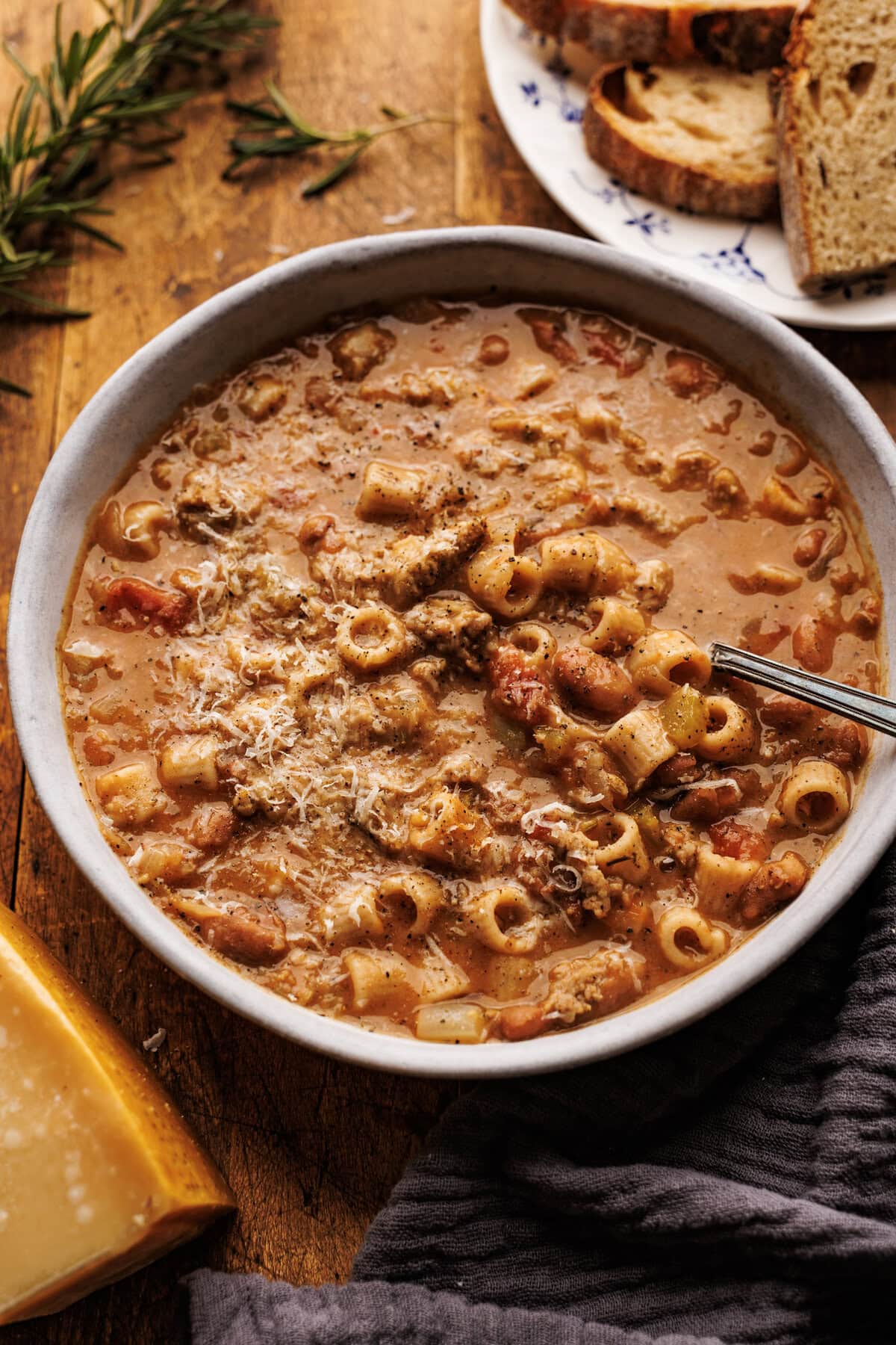A close up of pasta fagioli with sausage in a white bowl. Surrounding the bowl is a plate of bread slices, a block of Parmigiano Reggiano cheese, and a dark napkin. Everything is arranged on a wooden board.
