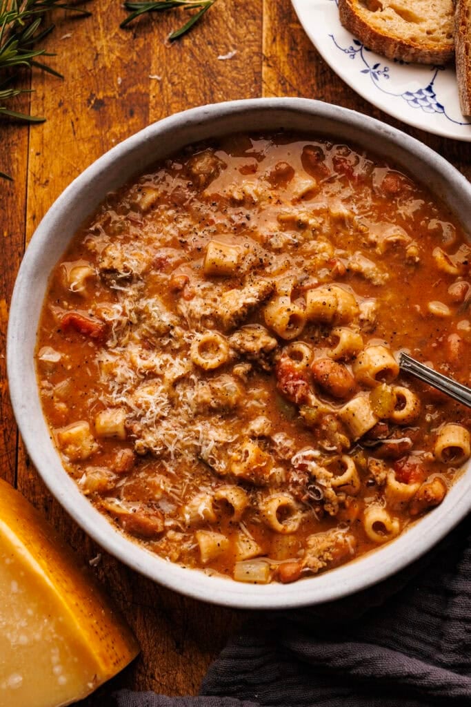 A close up of pasta fagioli with sausage in a white bowl. Surrounding the bowl is a plate of bread slices, a block of Parmigiano Reggiano cheese, and a dark napkin. Everything is arranged on a wooden board.