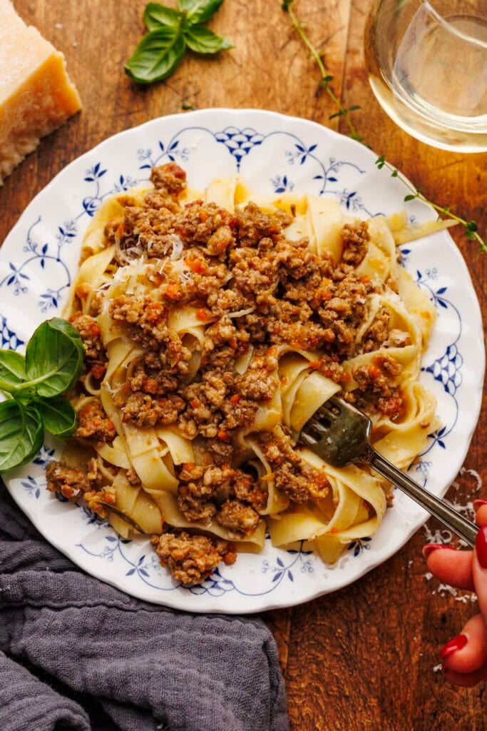 A white plate with white bolognese and tagliatelle pasta with a fork twirled in it. The plate of pasta is decorated on a wooden background with a glass of white wine, fresh basil leaves, and a dark gray napkin placed around it.