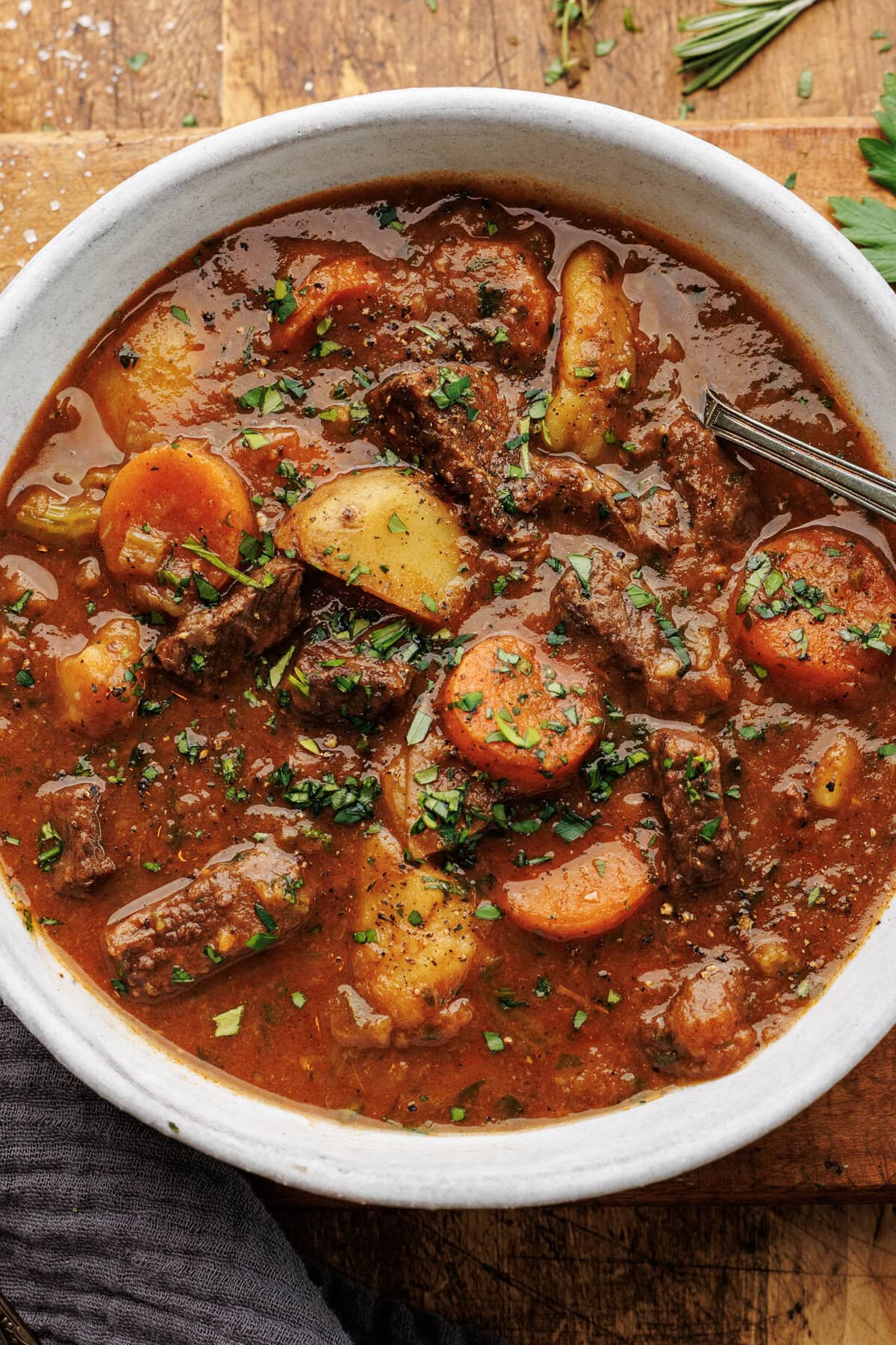 A close up of beef stew in a white bowl placed on a wooden board with herbs and a dark napkin decorated around it.